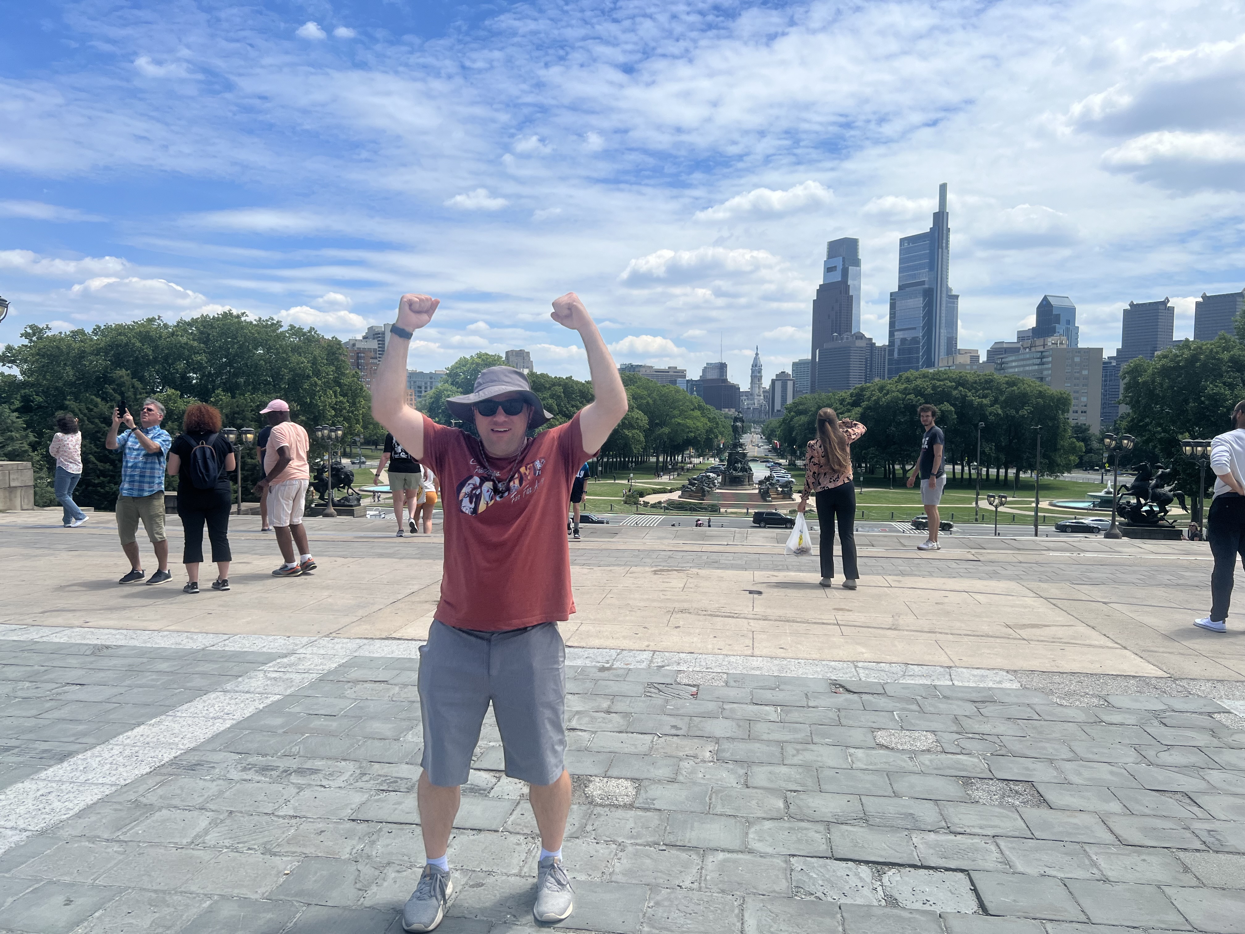 Brian raising both arms in a Rocky pose on the Philadelphia Museum of Art steps with the Philadelphia skyline behind him