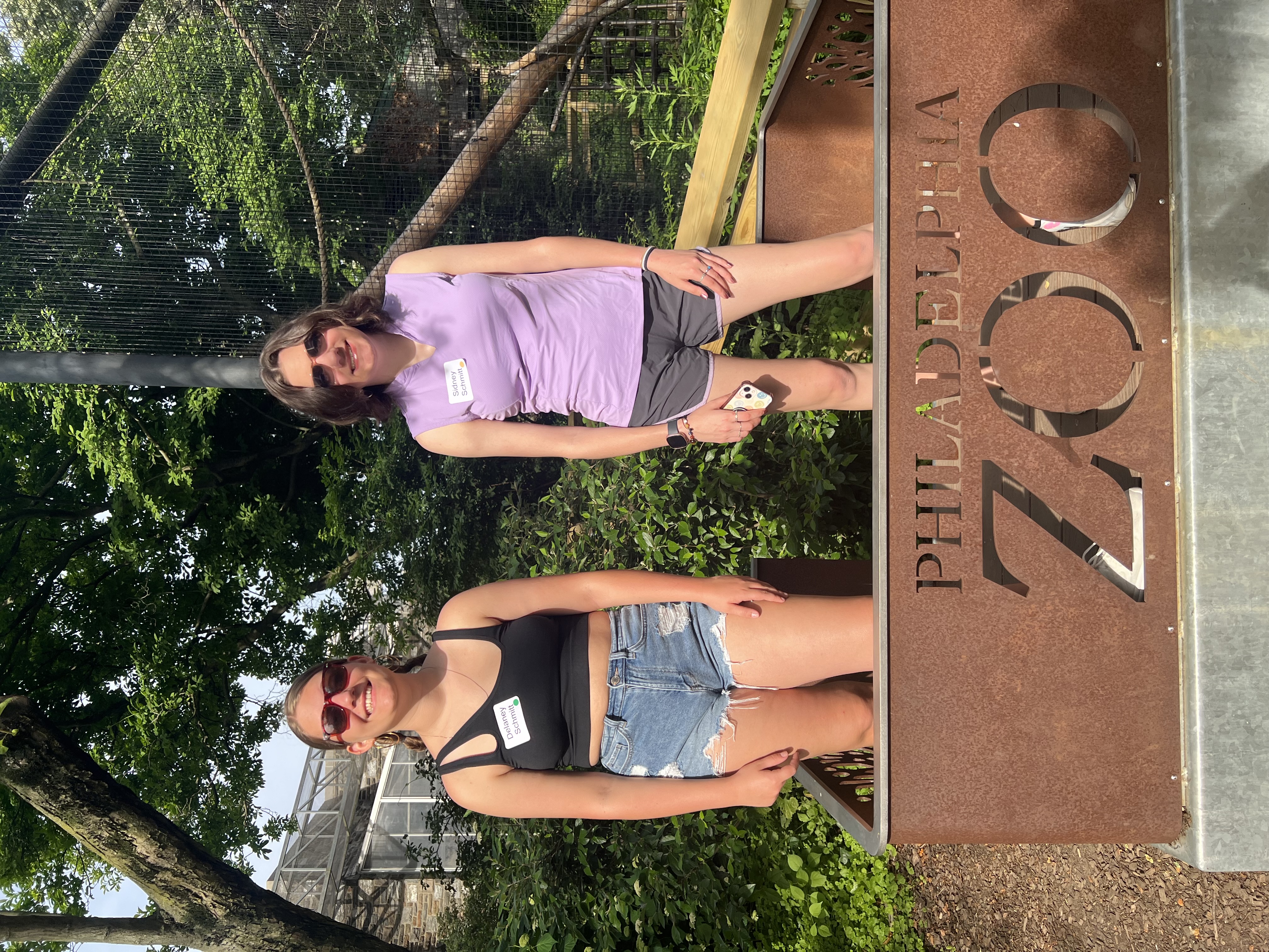 Two college-aged sisters standing beside the Philadelphia Zoo sign on a sunny day