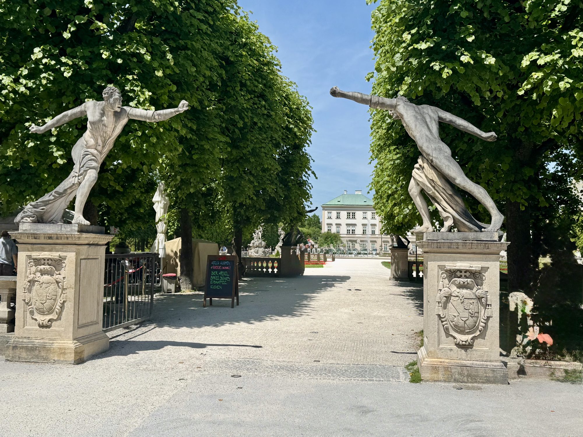Two stone statues framing the entrance to Mirabell Garden, with Mirabell Palace in the distance