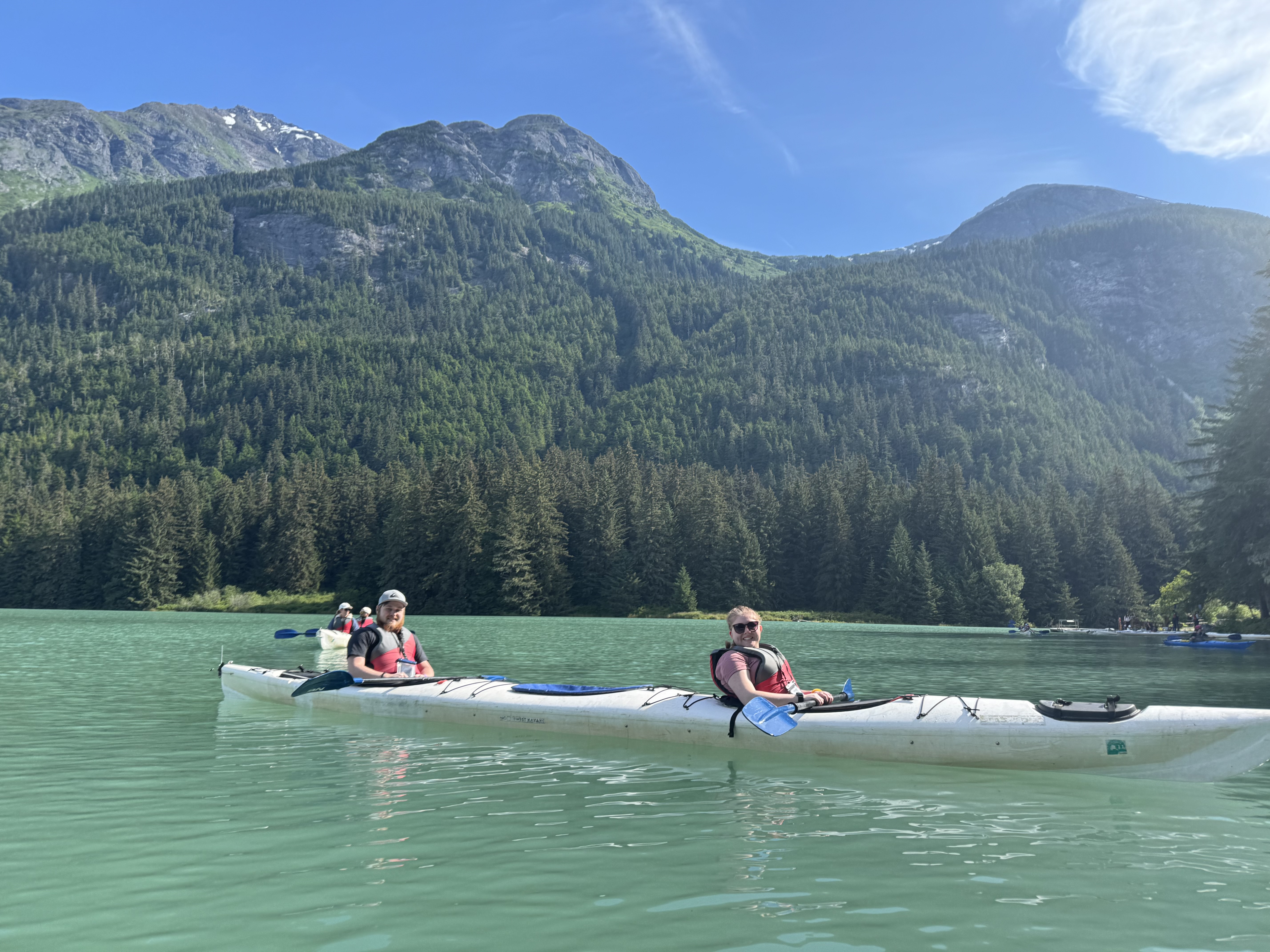 Two family members kayaking on turquoise Chilkoot Lake beneath forested mountains