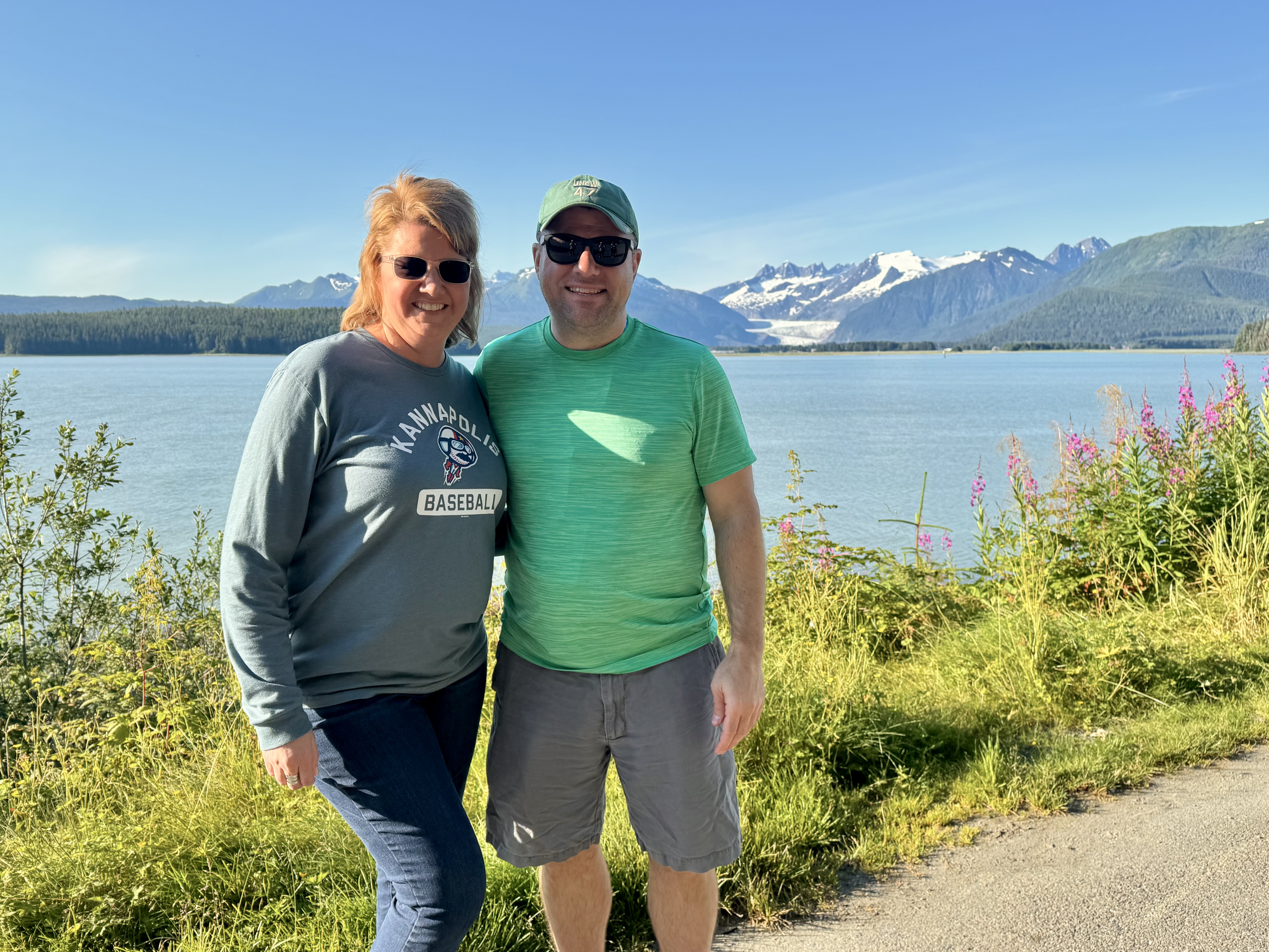 Angela and Brian standing beside a blue-green Alaska waterway with snowy mountains behind them