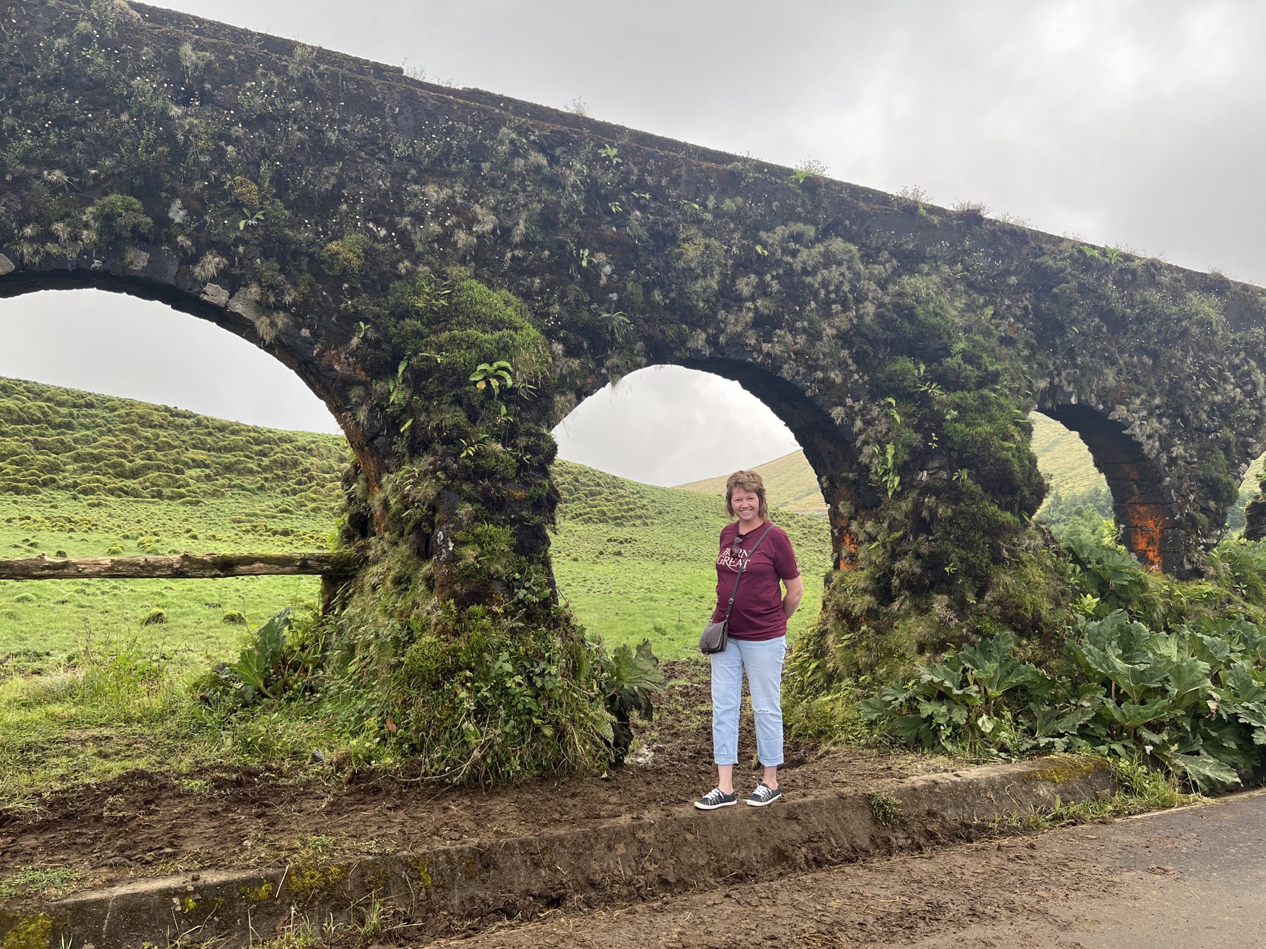 Angela standing beside a small moss-covered stone aqueduct in a green field