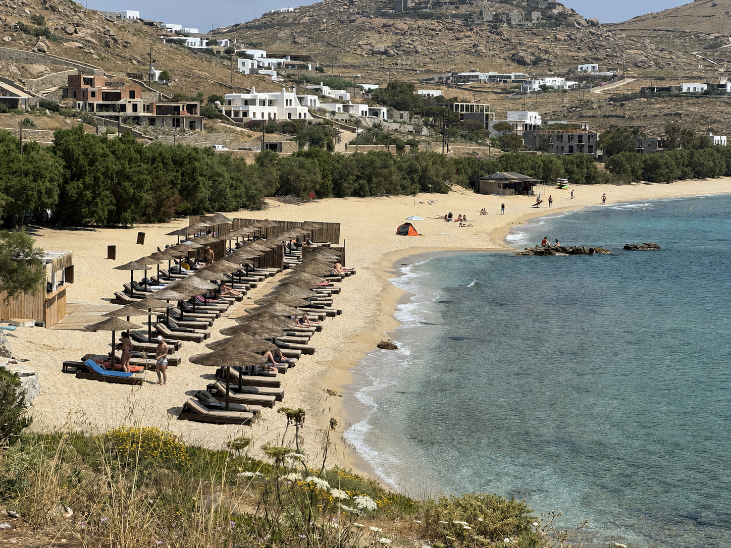A beach with rows of shaded loungers and white hillside buildings in Mykonos, Greece