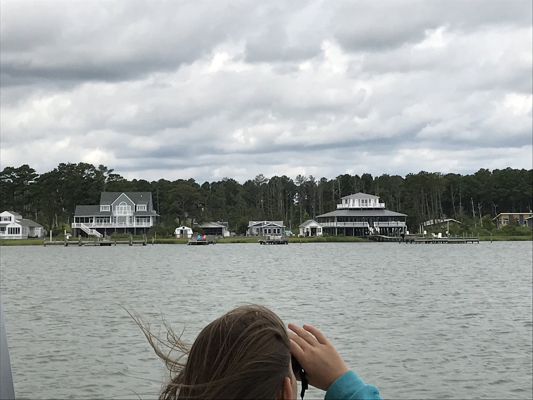 A child using binoculars from a boat while looking across the water toward shoreline homes