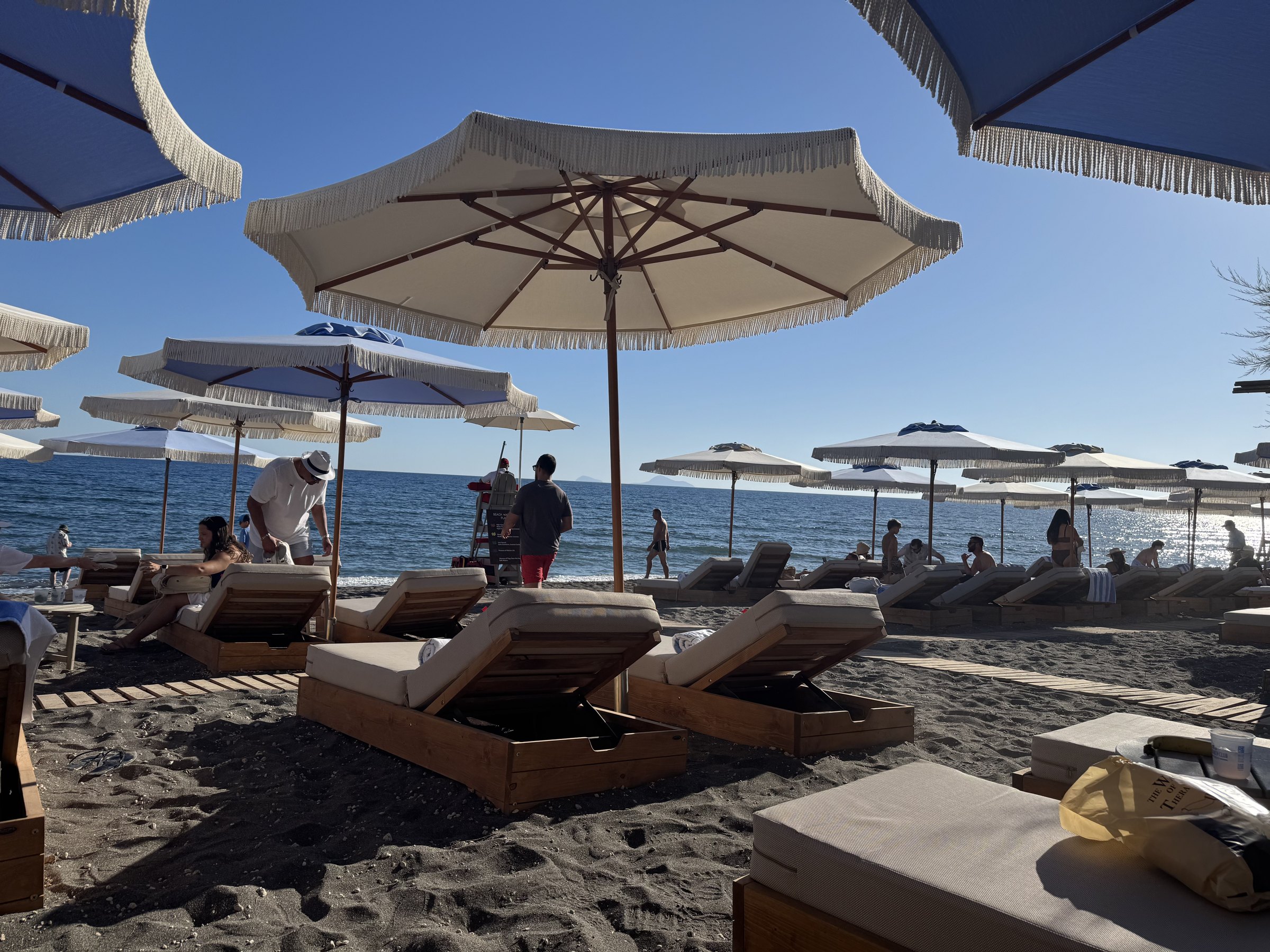 Loungers and fringed umbrellas arranged on the black-sand beach at Royal Beach Club Santorini