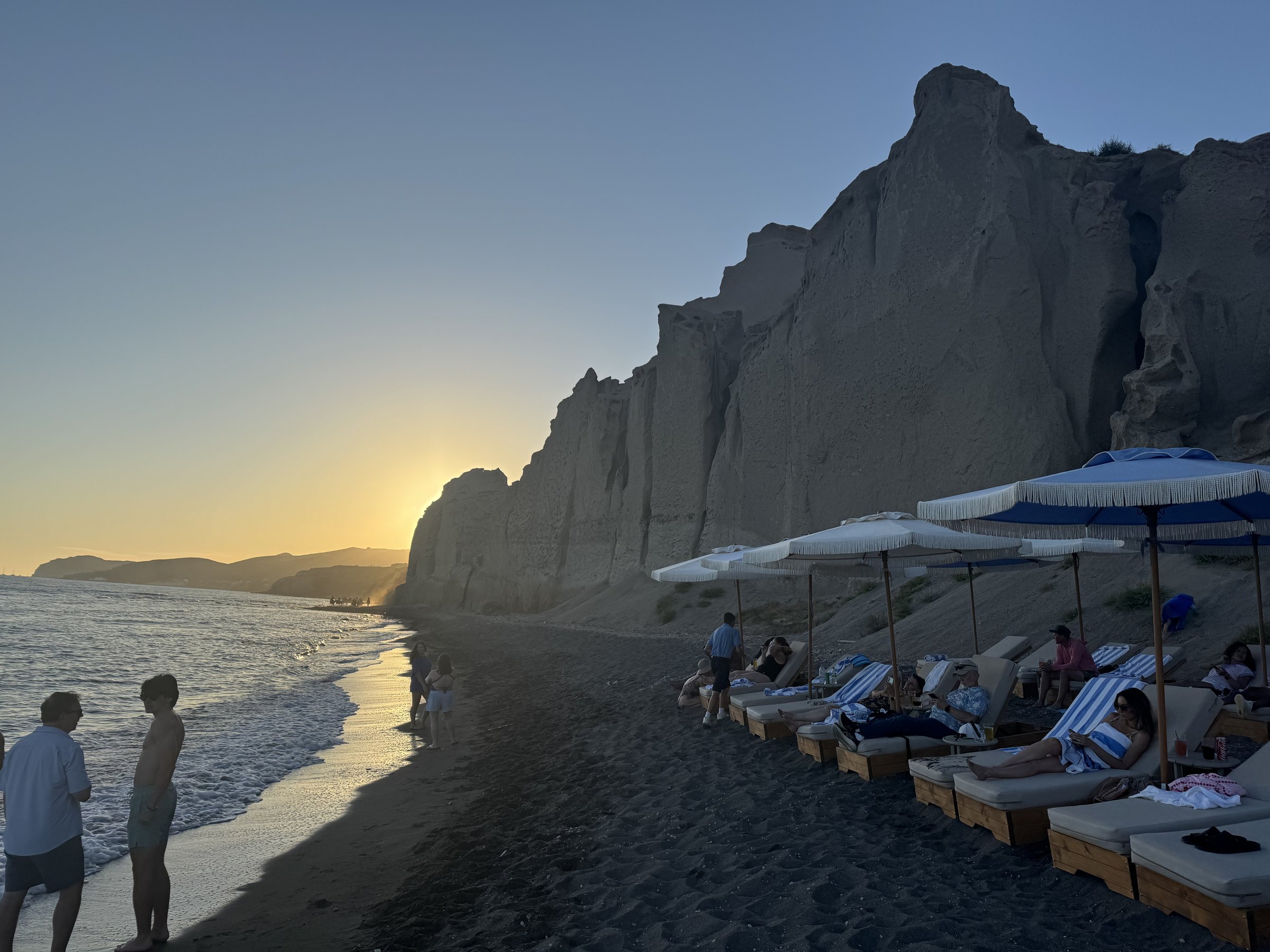 Guests relaxing on loungers beneath fringed umbrellas below cliffs at sunset