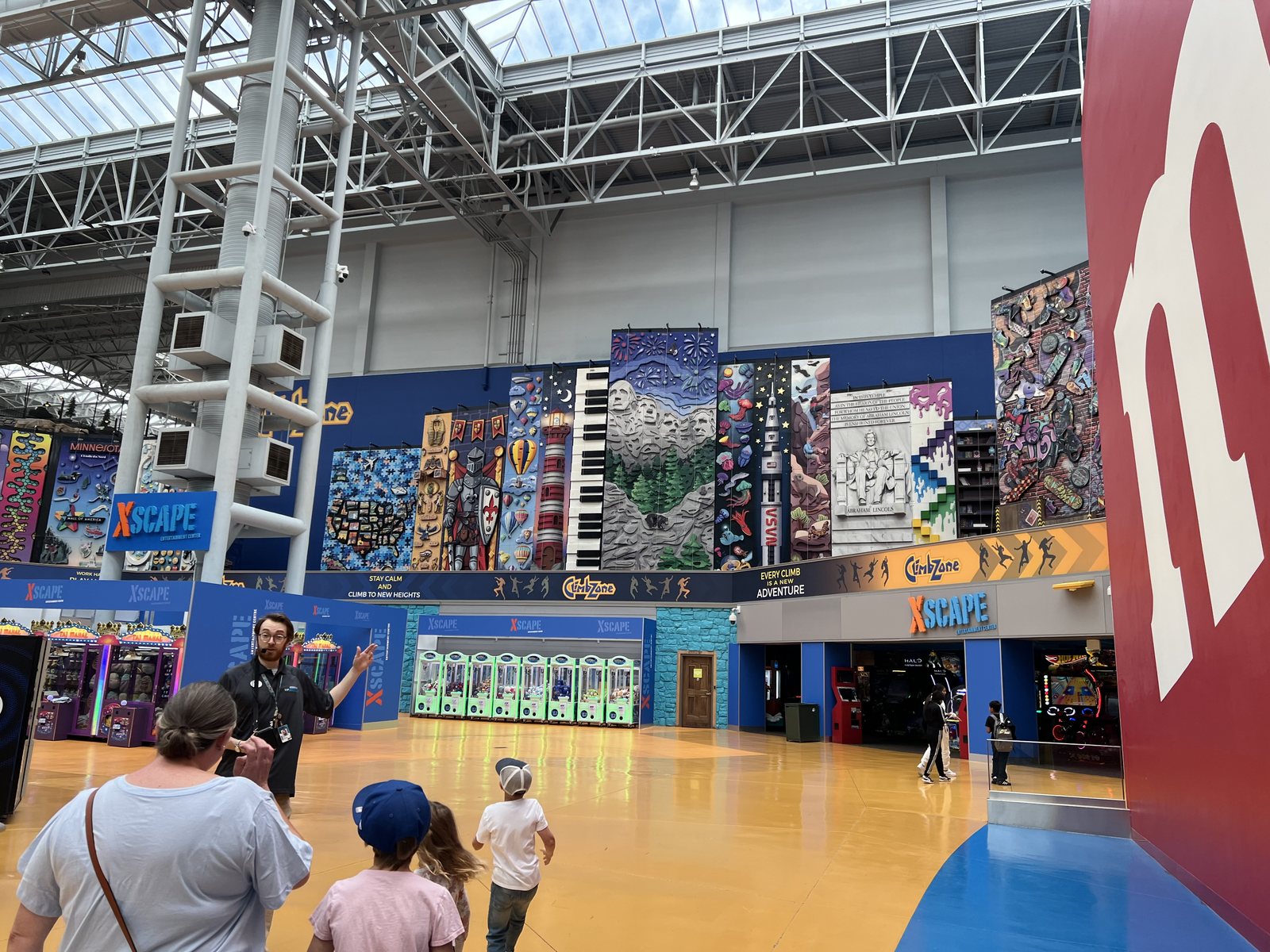 A Mall of America tour guide speaking beside colorful attractions and a Minnesota-themed climbing wall