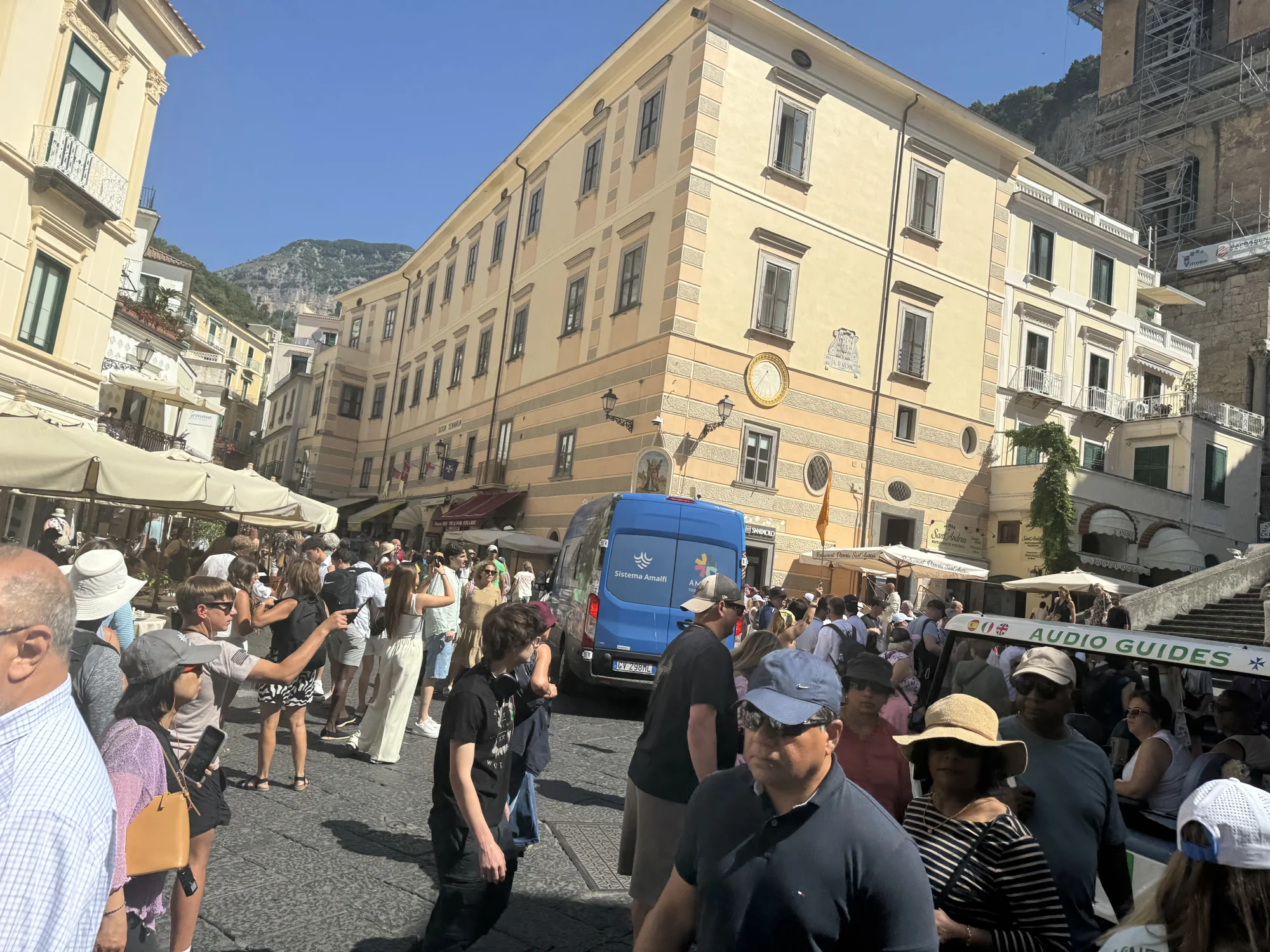 Crowds of visitors sharing a narrow passage with a blue van between Amalfi’s pastel buildings and stone steps