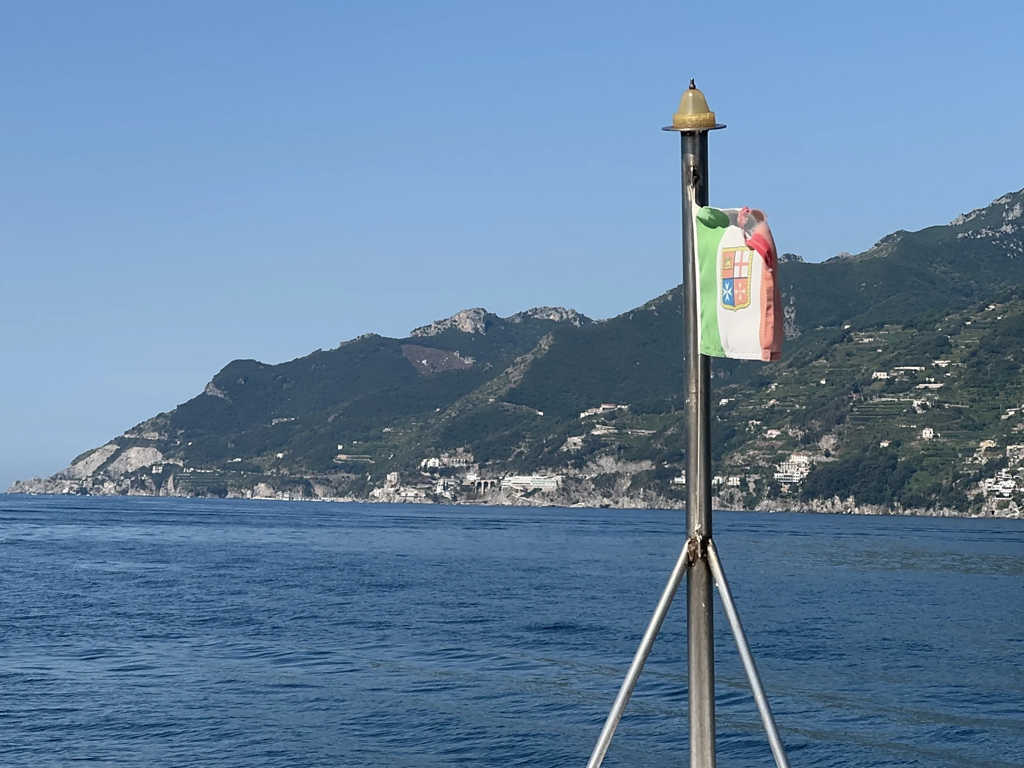 An Italian flag on the ferry with the mountainous coastline beyond blue water