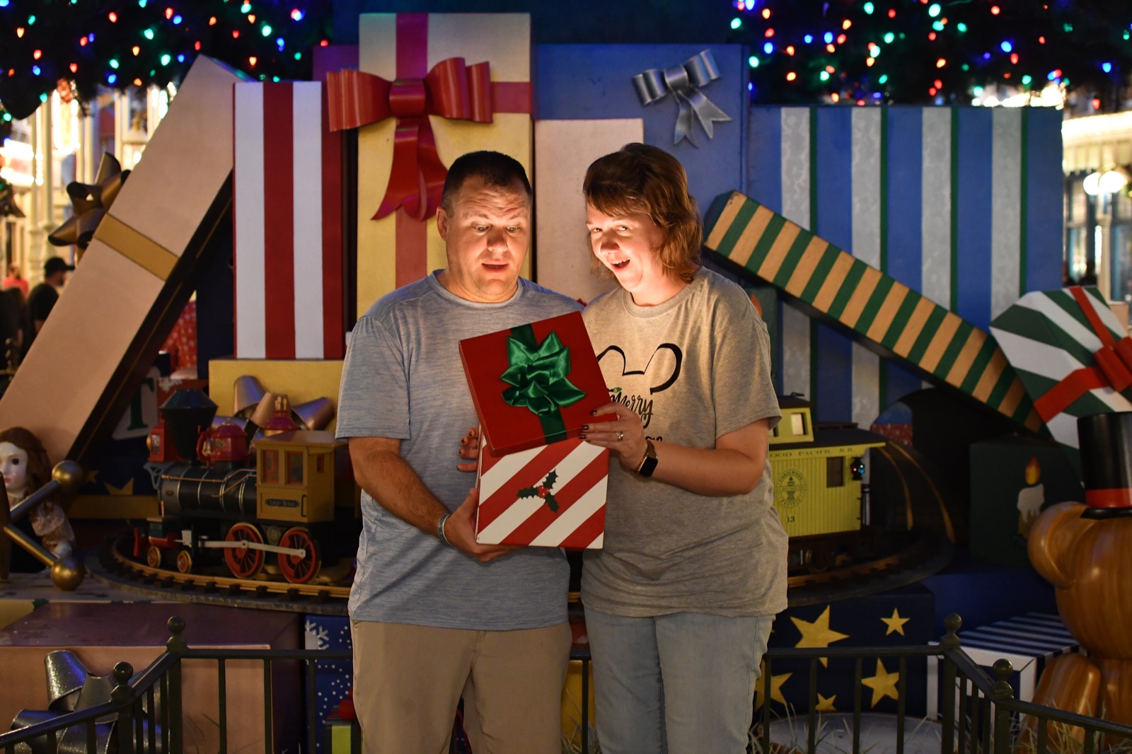 Angela and Brian opening a prop Christmas gift in front of oversized presents and holiday lights at Walt Disney World