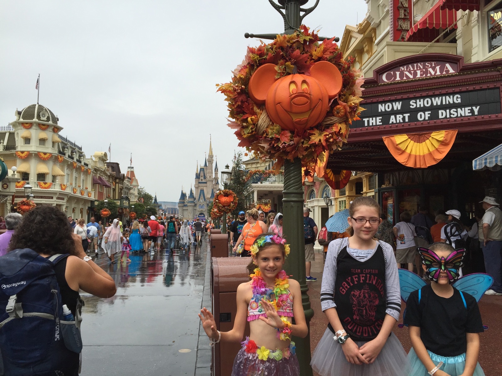 Angela’s three daughters in costumes beside a Mickey pumpkin wreath on a rain-wet Main Street, U.S.A.