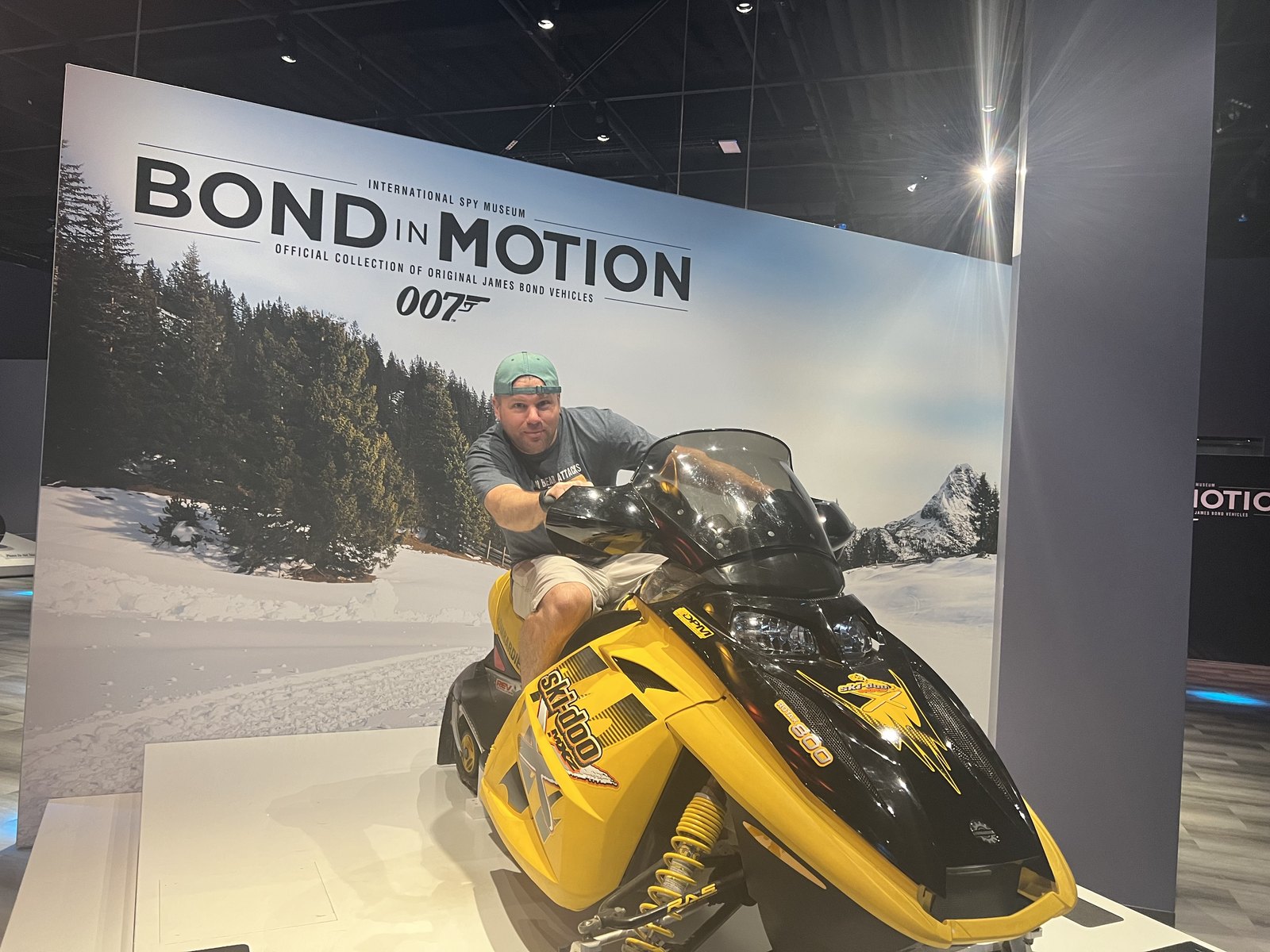 Brian posing on a yellow snowmobile in front of the Bond in Motion James Bond exhibition sign at the International Spy Museum
