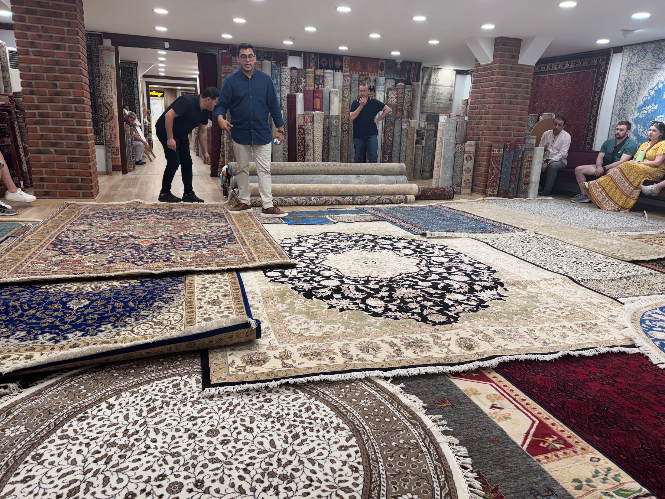 A presenter surrounded by Turkish carpets spread across the floor and stacked throughout a rug showroom near Selçuk